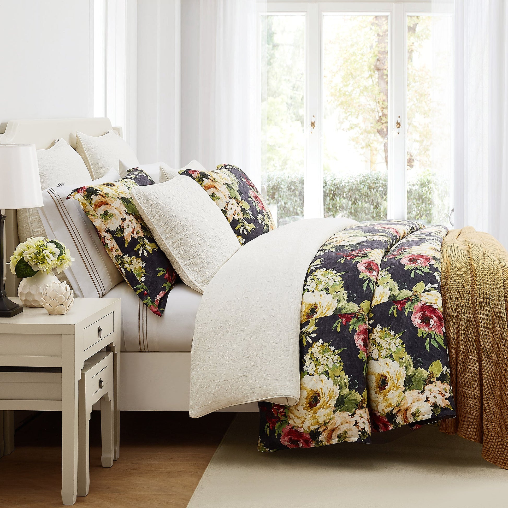 Floral comforter on a bed in a bright bedroom with a side table and lamp.